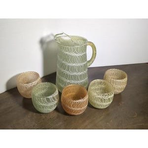 May include: A glass pitcher and five matching glasses. The pitcher and glasses are a pale green color with a white, web-like design. The glasses are round and the pitcher has a handle and a spout.