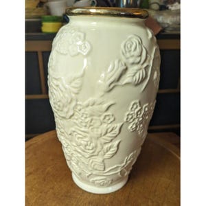 May include: Cream-colored ceramic vase with embossed floral designs, including roses and other flowers. The vase has a gold-colored rim and a rounded shape. The vase is sitting on a wooden surface.