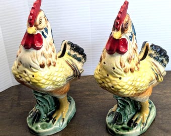 May include: Two ceramic rooster figurines. The roosters have yellow, brown, and cream-colored bodies with red combs and wattles. They stand on green bases and have black and blue accents. The figurines are likely decorative items.