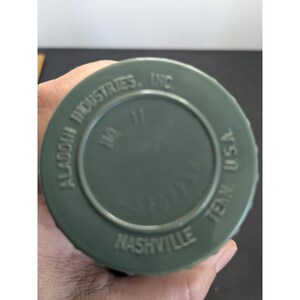 May include: A circular olive green plastic bottle cap with molded text reading ALADDIN INDUSTRIES, INC. NO. 11 NASHVILLE TENN. U.S.A. The cap features a ridged edge for grip and a central circular indentation, displaying clear manufacturing details.