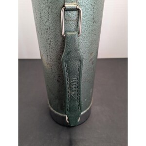 May include: A cylindrical metal thermos with a textured, dark green finish and a metallic base. A dark green leather-like strap is attached to the side with silver-toned metal hardware. The word Aladdin is embossed vertically on the strap.