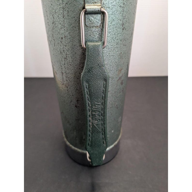 May include: A cylindrical metal thermos with a textured, dark green finish and a metallic base. A dark green leather-like strap is attached to the side with silver-toned metal hardware. The word Aladdin is embossed vertically on the strap.