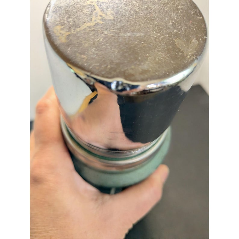 May include: A cylindrical vacuum insulated bottle featuring a textured sage green body and a polished chrome cap. The top of the cap has a metallic finish with visible surface wear and minor scratches, while the base of the cap is reflective.