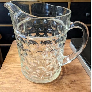 May include: A clear glass pitcher with a handle and a spout. The pitcher has a textured pattern of raised circles around the body. The pitcher is sitting on a wooden surface.