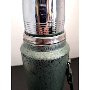 May include: A vintage-style vacuum flask featuring a textured, sage green metal exterior with visible signs of wear and a weathered finish. The top section consists of a polished, reflective chrome cap with horizontal engraved lines and a dark green ring.