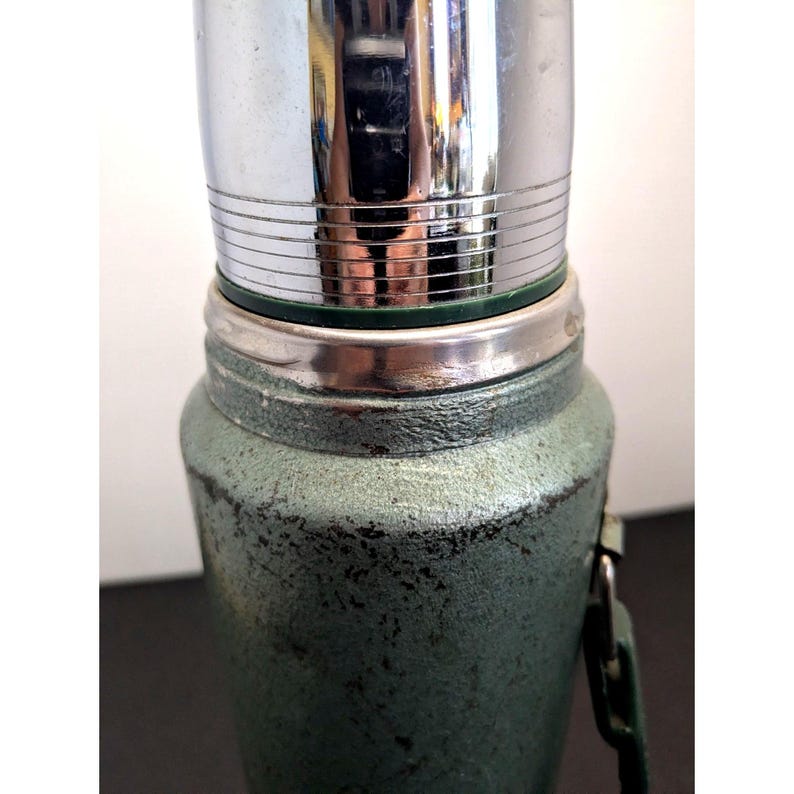May include: A vintage-style vacuum flask featuring a textured, sage green metal exterior with visible signs of wear and a weathered finish. The top section consists of a polished, reflective chrome cap with horizontal engraved lines and a dark green ring.