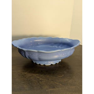 May include: A light blue ceramic bowl with a scalloped rim and decorative floral accents. The bowl has a pedestal base and a wide, shallow design. The bowl is likely used for serving or display.