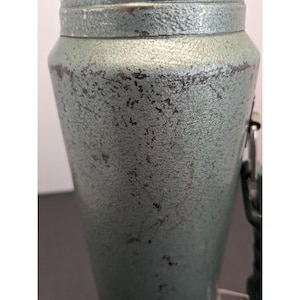 May include: A cylindrical metal container with a textured, sage green finish showing signs of wear and dark patina. The object features a metallic rim at the top and a side-mounted metal attachment point with a hinge, displaying a weathered surface.