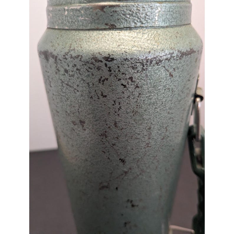 May include: A cylindrical metal container with a textured, sage green finish showing signs of wear and dark patina. The object features a metallic rim at the top and a side-mounted metal attachment point with a hinge, displaying a weathered surface.