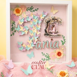 May include: A white shadow box frame containing a layered paper art display. The letter J is formed by pastel-colored paper butterflies. Next to it is a miniature illustration of a girl on a swing under a tree. The name Tania is written in script.