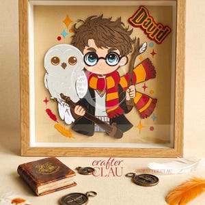 Peut inclure: Un cadre carré en bois contenant une œuvre d'art en papier superposé représentant un personnage de dessin animé aux cheveux bruns, lunettes rondes et écharpe rouge et jaune, tenant une baguette près d'un hibou blanc. Le nom David est inscrit.