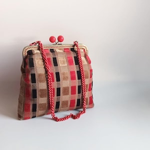 Può includere: Una borsa in stile vintage con un motivo geometrico a quadrati rossi, neri e beige. La borsa presenta una chiusura dorata con chiusure a sfera rosse e una tracolla in corda rossa abbinata.