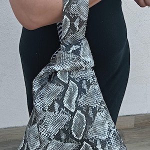 Könnte beinhalten: Eine dreieckige Handtasche mit Schlangenmuster in Grau- und Weißtönen. Die Tasche hat einen geknoteten Griff. Die Tasche wird von einer Person getragen, die ein schwarzes Kleid trägt. Das Design der Tasche ist auffällig und stilvoll.