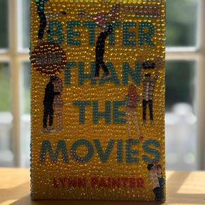 May include: A hardback book titled "Better Than The Movies" by Lynn Painter. The cover is yellow and covered in sparkling embellishments. The title is in teal letters, and there are illustrations of people.
