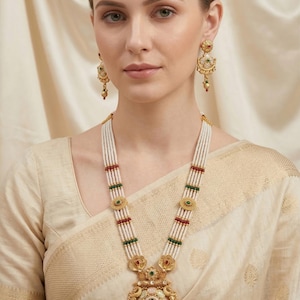 May include: A gold necklace and matching earrings set. The necklace has multiple strands of white beads, with red, green, and gold accents. The pendant features detailed patterns and colourful gemstones.