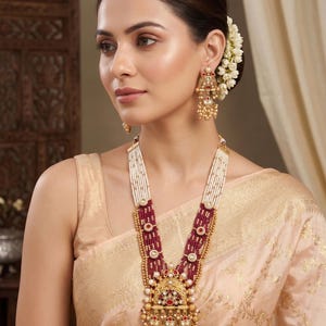 May include: A gold-toned necklace and matching earrings set. The necklace features a multi-strand design with white beads, burgundy beads, and gold accents. The pendant and earrings have intricate detailing with red and pearl-like embellishments.