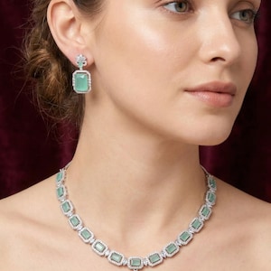 Puede incluir: Un conjunto de joyas a juego con un collar y pendientes. El collar y los pendientes tienen un engaste plateado con una serie de piedras rectangulares de color verde claro. Las piedras están rodeadas de piedras más pequeñas y transparentes.