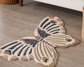 Puede incluir: Una alfombra con forma de mariposa en beige, rosa y azul marino. La alfombra presenta un diseño detallado de alas con un borde marrón. Se encuentra sobre un suelo de madera cerca de un sofá blanco.