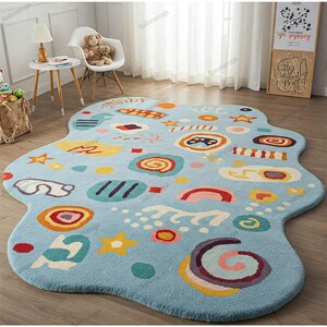 May include: A light blue, irregularly shaped rug with colorful, whimsical designs. The rug features various shapes like stars, circles, and cloud-like forms in shades of yellow, orange, red, and purple. The rug is placed on a wooden floor.