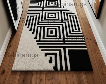 May include: A rectangular rug with a geometric black and white design. The rug features a repeating pattern of squares and lines, with a solid black section at one end. The rug is placed on a wooden floor.