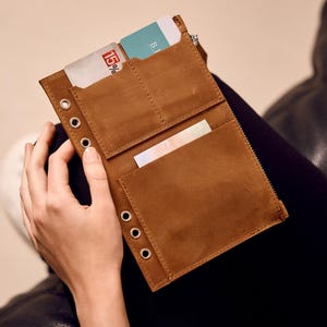 May include: A brown leather wallet featuring multiple card slots, a zippered compartment, and a vertical row of five silver-toned metal eyelets along the left edge. The wallet contains two visible cards, one displaying the number 15 percent.