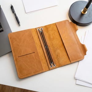 May include: A tan leather traveler's notebook cover featuring a smooth exterior, a central spine with two black elastic cords secured by metal eyelets, and interior pockets on both the left and right sides for holding inserts.