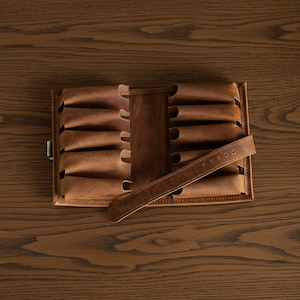 Puede incluir: Un estuche de cuero marrón para puros, abierto y plano. El estuche tiene múltiples ranuras para puros y un cierre de correa de cuero. El cuero tiene un aspecto envejecido, con variaciones de color.