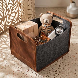 Könnte beinhalten: Eine Aufbewahrungsbox mit brauner Lederaußenseite und grauem Filzinnenraum. Die Box enthält einen Teddybären mit gestreiftem Schal, Holzblöcke, ein Spielzeugauto und Mikadospiele. Die Box hat zwei Griffe.