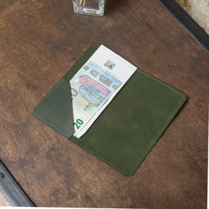Puede incluir: Una cartera de cuero verde oliva con un billete de veinte euros parcialmente visible. La cartera tiene un diseño sencillo con un bolsillo verde oscuro en contraste. La cartera rectangular parece ser de ante. Dimensiones: aproximadamente 15 cm x 10 cm.