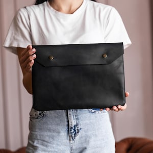 Könnte beinhalten: Schwarze Laptop-Hülle aus Leder mit zwei messingfarbenen Druckknöpfen. Die Hülle ist rechteckig und hat eine Klappe, die sich über die Oberseite faltet. Das Leder scheint glatt zu sein und das Design ist minimalistisch.