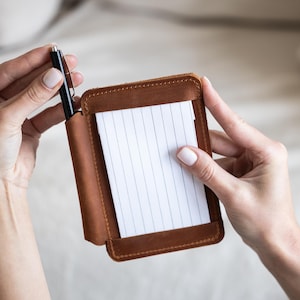 May include: A brown leather notepad holder with a pen slot and a notepad with lined paper. The notepad holder is rectangular and has a stitched border. A black pen is inserted into the pen slot.