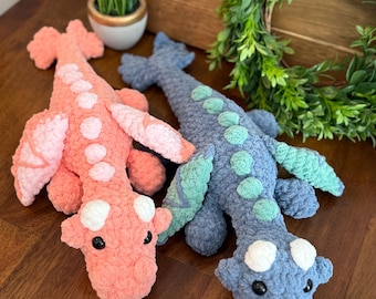 May include: Two handmade crochet dragon plushies. One is coral with white spots and pink wings, the other is blue with green spots and wings. Both have black eyes and are lying on a wooden surface.