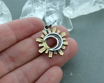 May include: A pendant featuring a sun and moon design. The pendant has a silver-colored crescent moon in the center, surrounded by a silver border and gold-colored sun rays. The pendant is held in a hand.