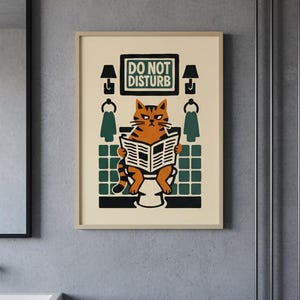 May include: A framed print featuring a cartoon cat reading a newspaper while sitting on a toilet. The cat is orange and black, with a grumpy expression. The print includes the text "DO NOT DISTURB" in a green and white box.