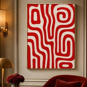 May include: A framed abstract art piece with a red and white pattern hangs above a red velvet armchair. The artwork features a bold, swirling design. A gold-toned lamp and a small side table with a vase of red flowers complete the scene.