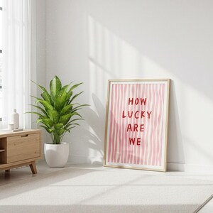 Op de afbeelding: Een ingelijste print met de tekst "HOW LUCKY ARE WE" in rood op een roze en witte gestreepte achtergrond. De print staat tegen een witte muur in een kamer met een plant en houten meubels.