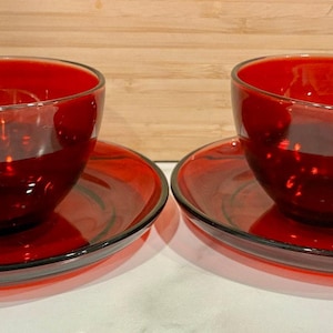 Puede incluir: Dos tazas de té de vidrio rojo a juego con platillos. Las tazas tienen forma redondeada con asa, y los platillos son circulares. El juego está hecho de vidrio rojo transparente, con un acabado brillante. Las tazas y los platillos están colocados sobre una superficie blanca.