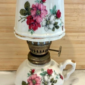 May include: A small, white ceramic oil lamp with a floral design featuring pink and red roses and white flowers. The lamp has a brass burner and a small handle. The lampshade is also white with the same floral pattern.