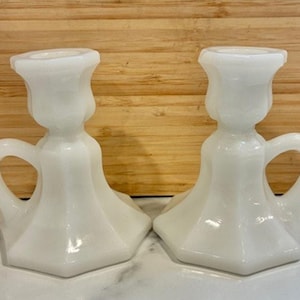 May include: A pair of white milk glass candle holders. Each has a flared base, a handle, and a cup for a candle. The candle holders are made of smooth, opaque white glass and are set against a wooden background.