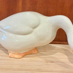 May include: A ceramic duck figurine in a glossy white finish. The duck has an orange beak and feet, with a black eye. The duck is in a downward-facing pose, as if it is looking down. The figurine is on a wooden surface.