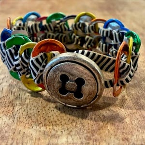 May include: A bracelet constructed from colorful metal soda can tabs woven with black and white striped fabric. The closure features a large, round, antique silver-toned button with four holes in the center, securing the circular band together.