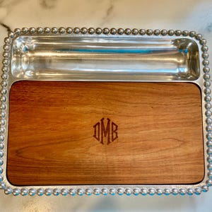 May include: A rectangular serving tray with a silver beaded border. The tray has a wooden base with a monogram and a silver compartment. The wooden section is a warm brown color.