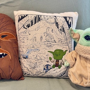 Puede incluir: Tres peluches con temática de Star Wars: una almohada de Chewbacca, una almohada con una ilustración en blanco y negro de Yoda y una escena, y un muñeco de peluche de Grogu. La almohada de Chewbacca es marrón con detalles en naranja.