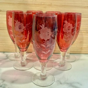 May include: A set of eight red champagne flutes. Each glass features a clear, etched floral design. The flutes have a long stem and a round base. The glasses are arranged on a white surface.
