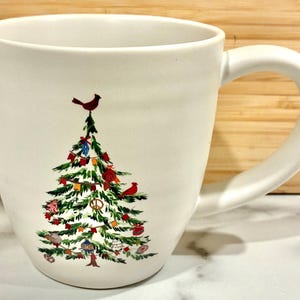 May include: White ceramic mug with a large handle, decorated with a Christmas tree design. The tree is adorned with ornaments and red birds. The mug is set against a light wood background.