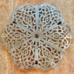 May include: A silver-colored, ornate metal coaster with a floral design. The coaster has a circular shape with a central hole and a scalloped edge. The intricate openwork pattern is visible against a wooden background.