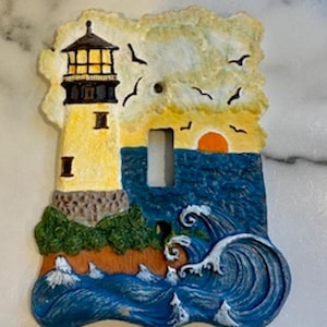 Puede incluir: Cubierta decorativa para interruptor de luz con un diseño de faro. La cubierta muestra un faro amarillo con la parte superior negra, sobre un océano azul con olas rompiendo y una puesta de sol. Incluye pájaros volando.