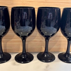 May include: Four black wine glasses with a rounded bowl and a long stem. The glasses are made of a shiny, reflective material and are arranged in a row. The glasses are on a light-colored wooden surface.