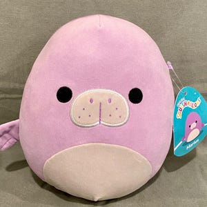 May include: A plush, lavender-colored seal with black eyes and a light beige snout and belly. The stuffed animal has a small fin and a tag that reads "Original Squishmallows" and the name "Marius".