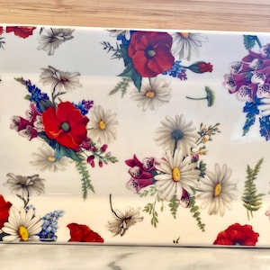 May include: Rectangular serving platter with a white background, adorned with a vibrant floral pattern. The design features red poppies, white daisies, purple foxgloves, and blue wildflowers. A decorative piece for serving food.
